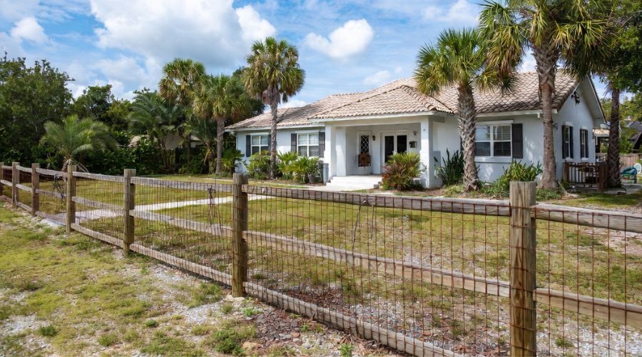 A white single-story house with a tan tiled roof surrounded by palm trees, enclosed by a wire fence with wooden posts.