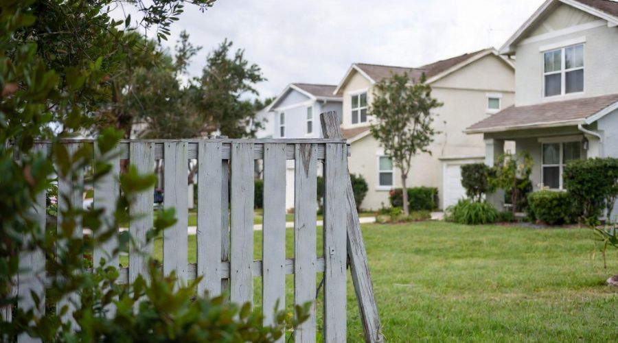 A wooden fence stands in the foreground, with a row of suburban houses and green lawns visible in the background.