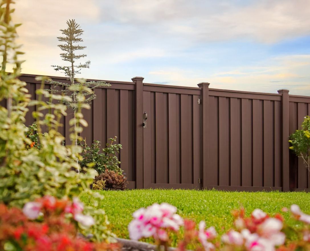A brown wooden fence with a gate in a backyard with flowers in the foreground.