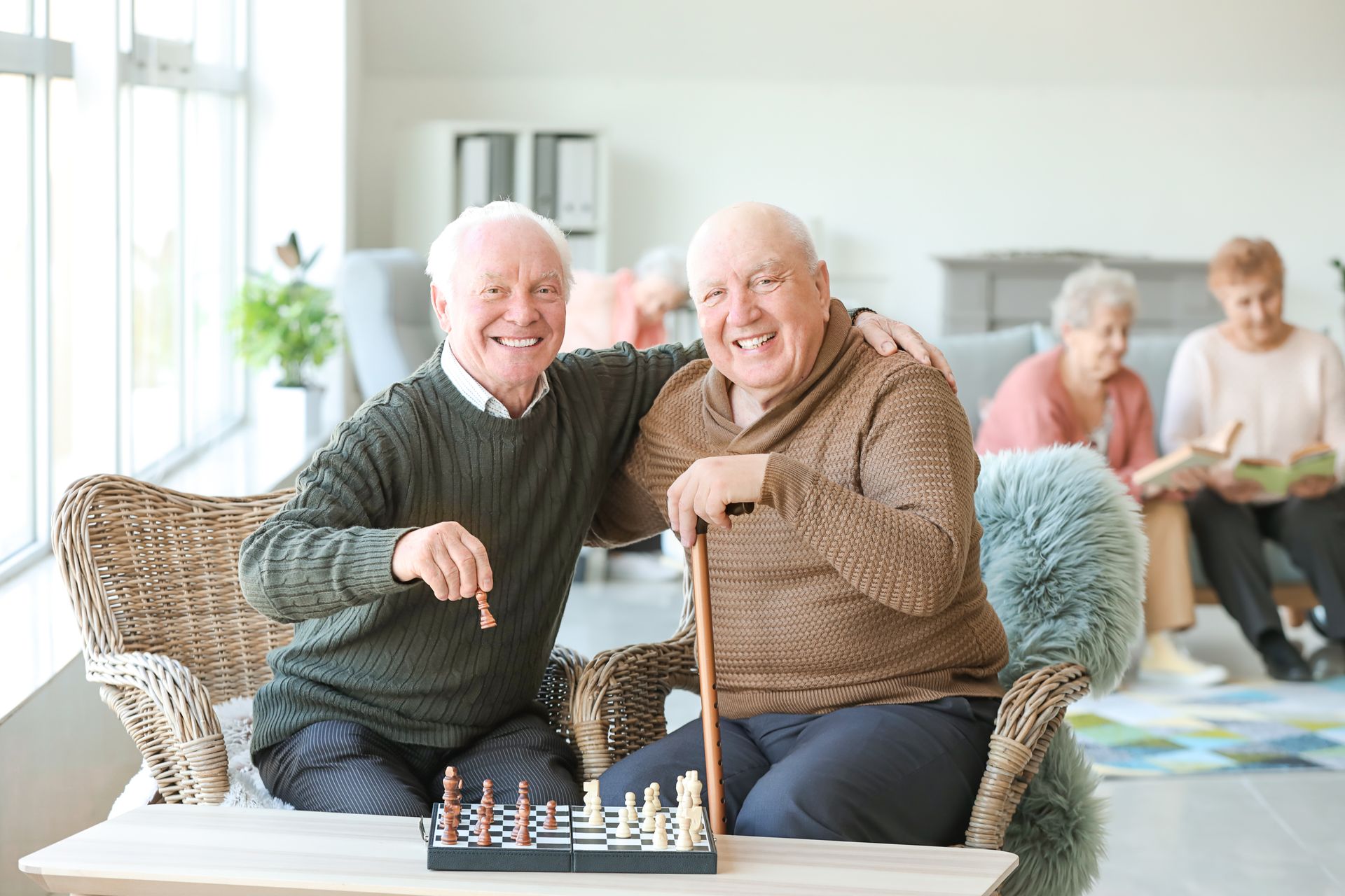 Two older men playing chess, smiling, one with arm around the other. Others in background. Two older men playing chess, smiling, one with arm around the other. Others in background.