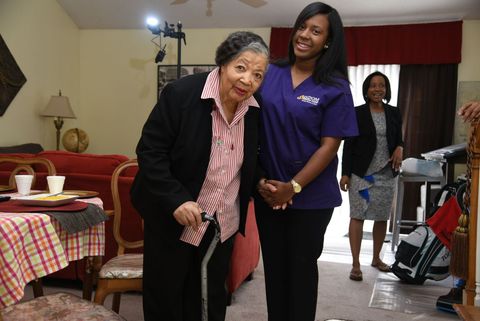 Caregiver helps elderly man walk with a cane, both smiling indoors. Caregiver helps elderly man walk with a cane, both smiling indoors.