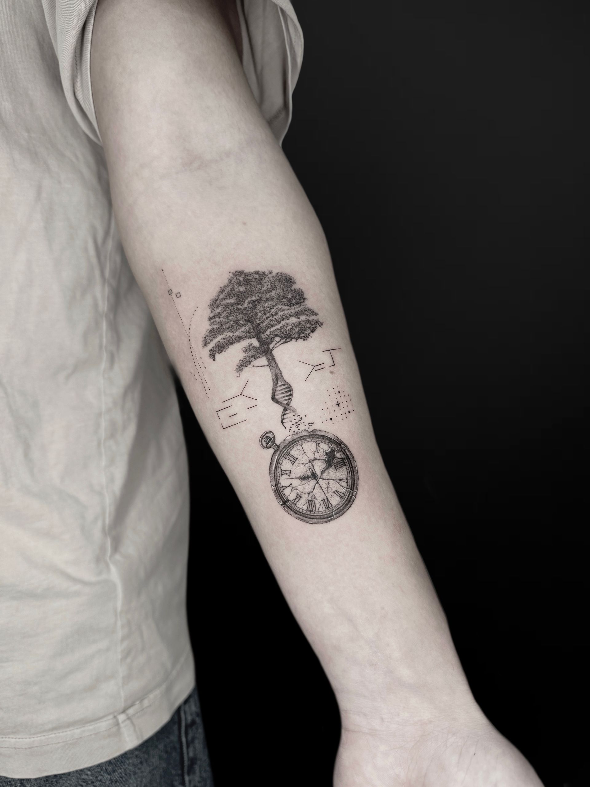 A black-and-grey tattoo on a forearm showing a tree whose roots transition into a DNA strand leading to a pocket watch.