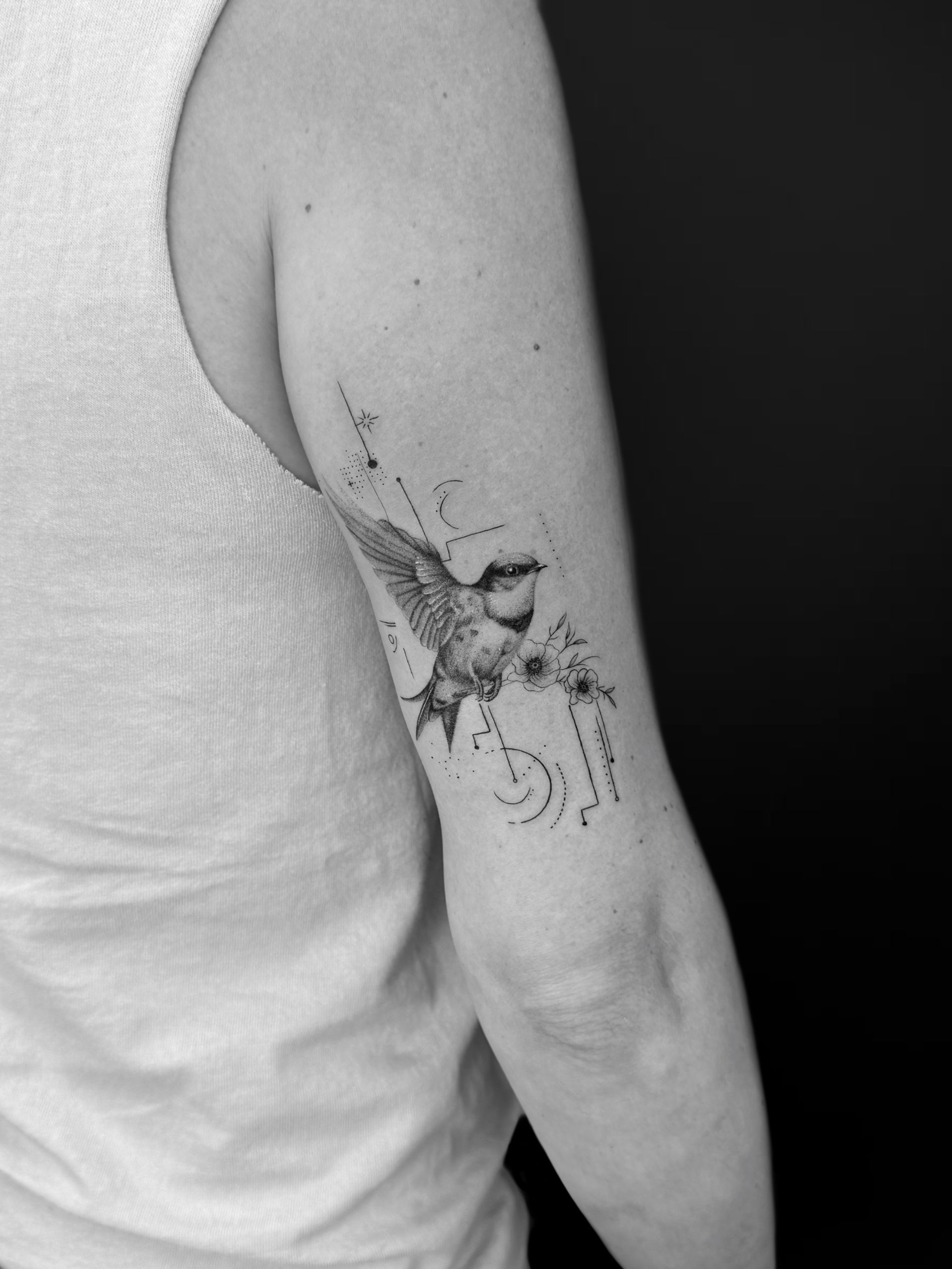 A black and white close-up of a bird tattoo with geometric, dot-work details on the back of an upper arm.