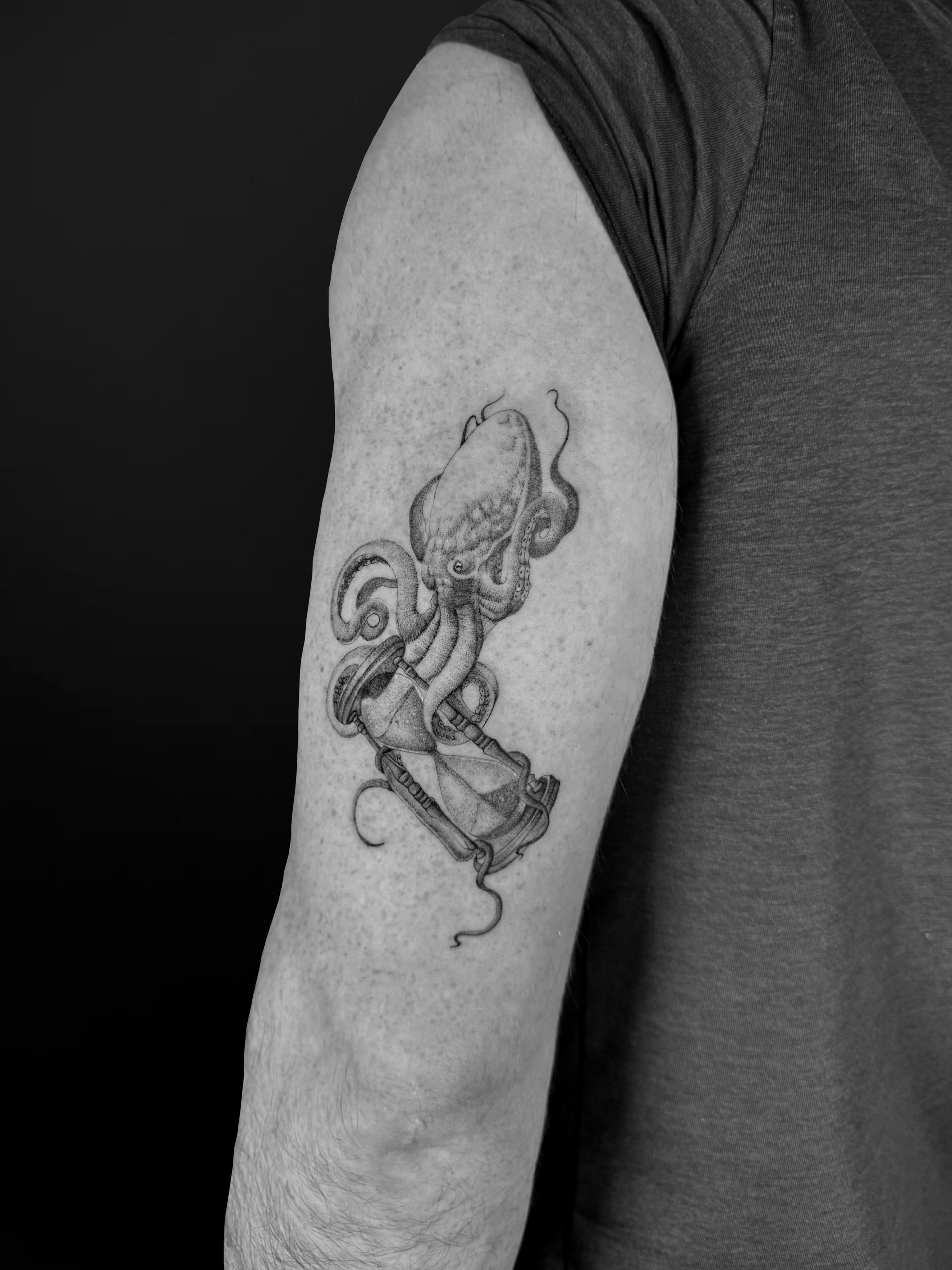 A black and white tattoo on an upper arm depicting an octopus wrapped around an hourglass.