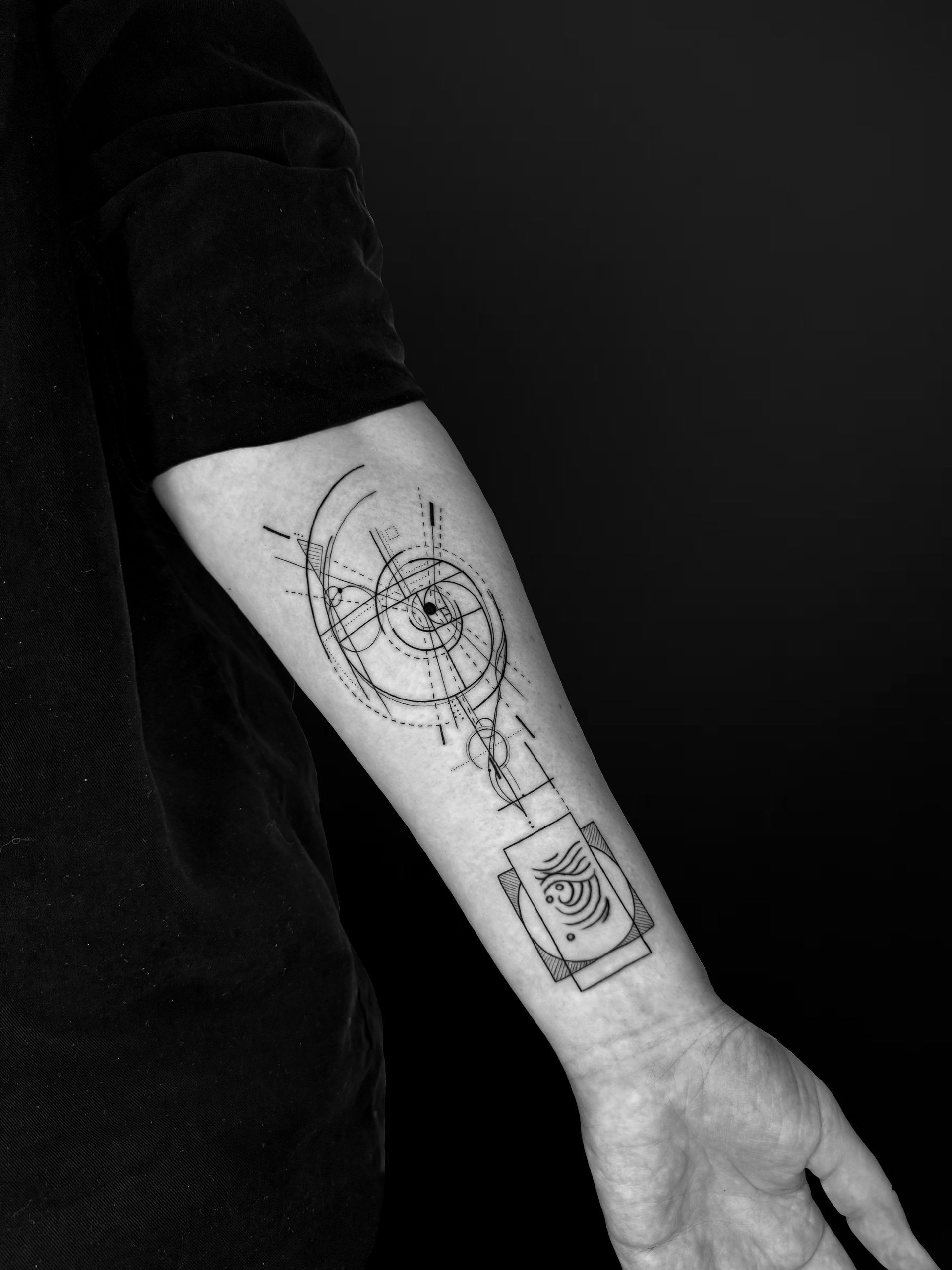 A forearm tattoo featuring delicate, thin-line geometric patterns, spiral orbits, and a rectangular architectural detail.