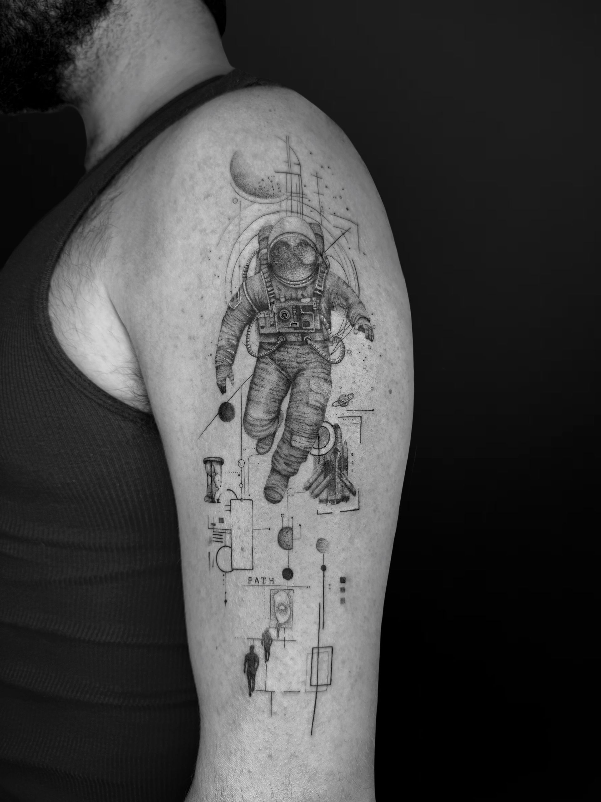 Black-and-grey tattoo on an upper arm featuring an astronaut floating among celestial bodies and abstract geometry.