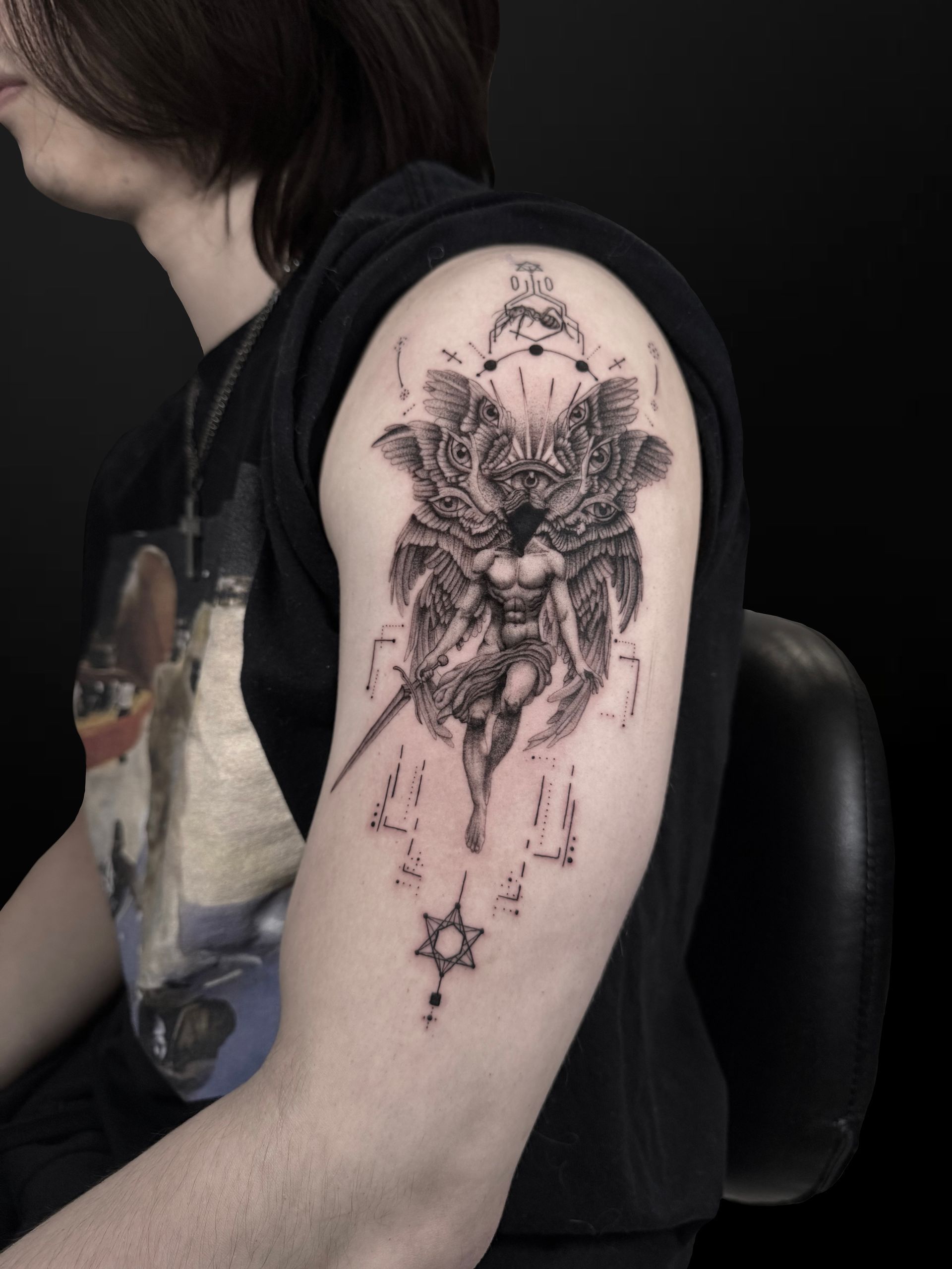 A detailed black-ink tattoo on an upper arm depicting a winged, angelic figure wielding a sword, with geometric symbols.