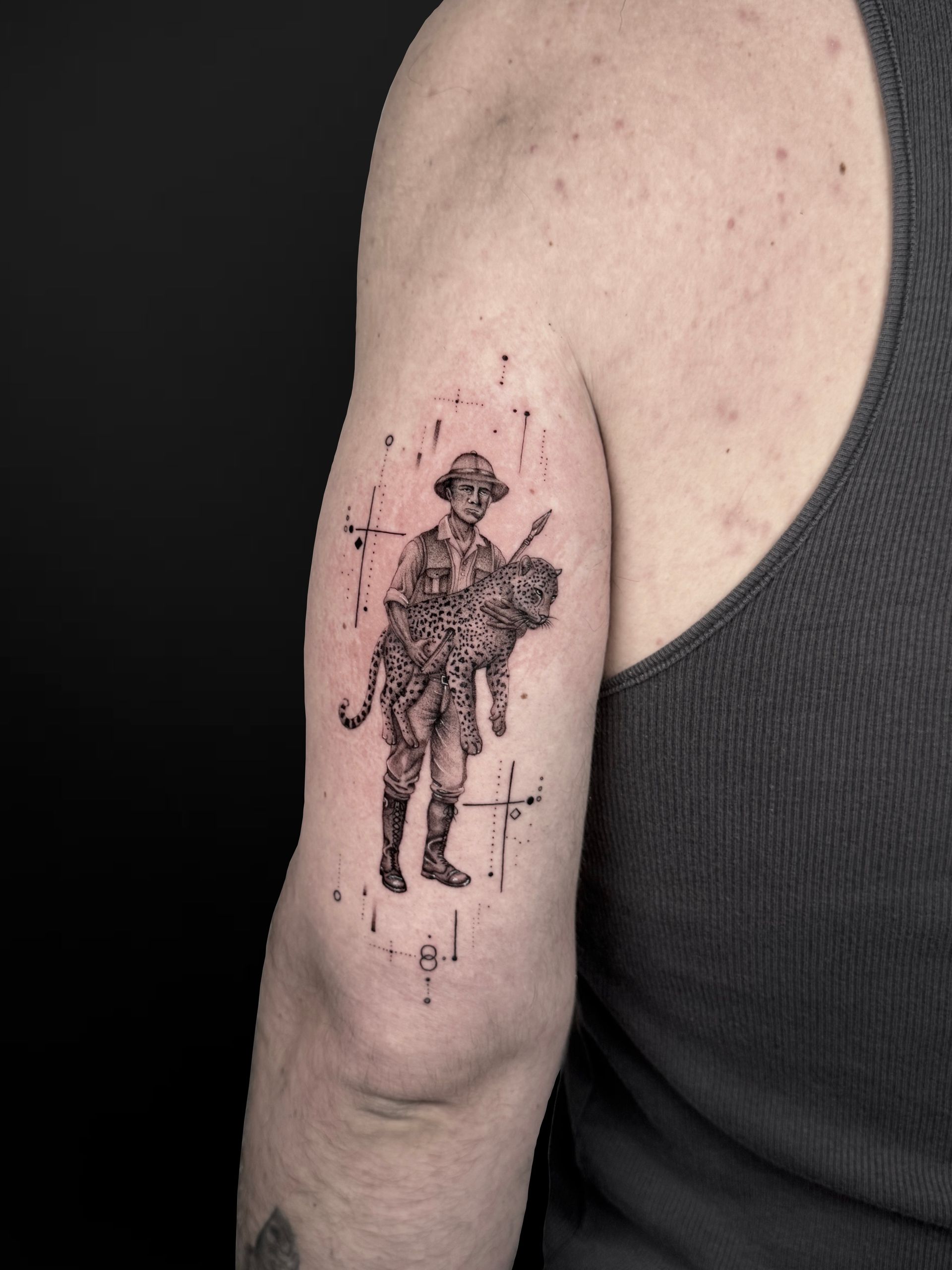 A detailed black-ink tattoo on an arm showing a person in explorer attire carrying a tiger cub, surrounded by star icons.
