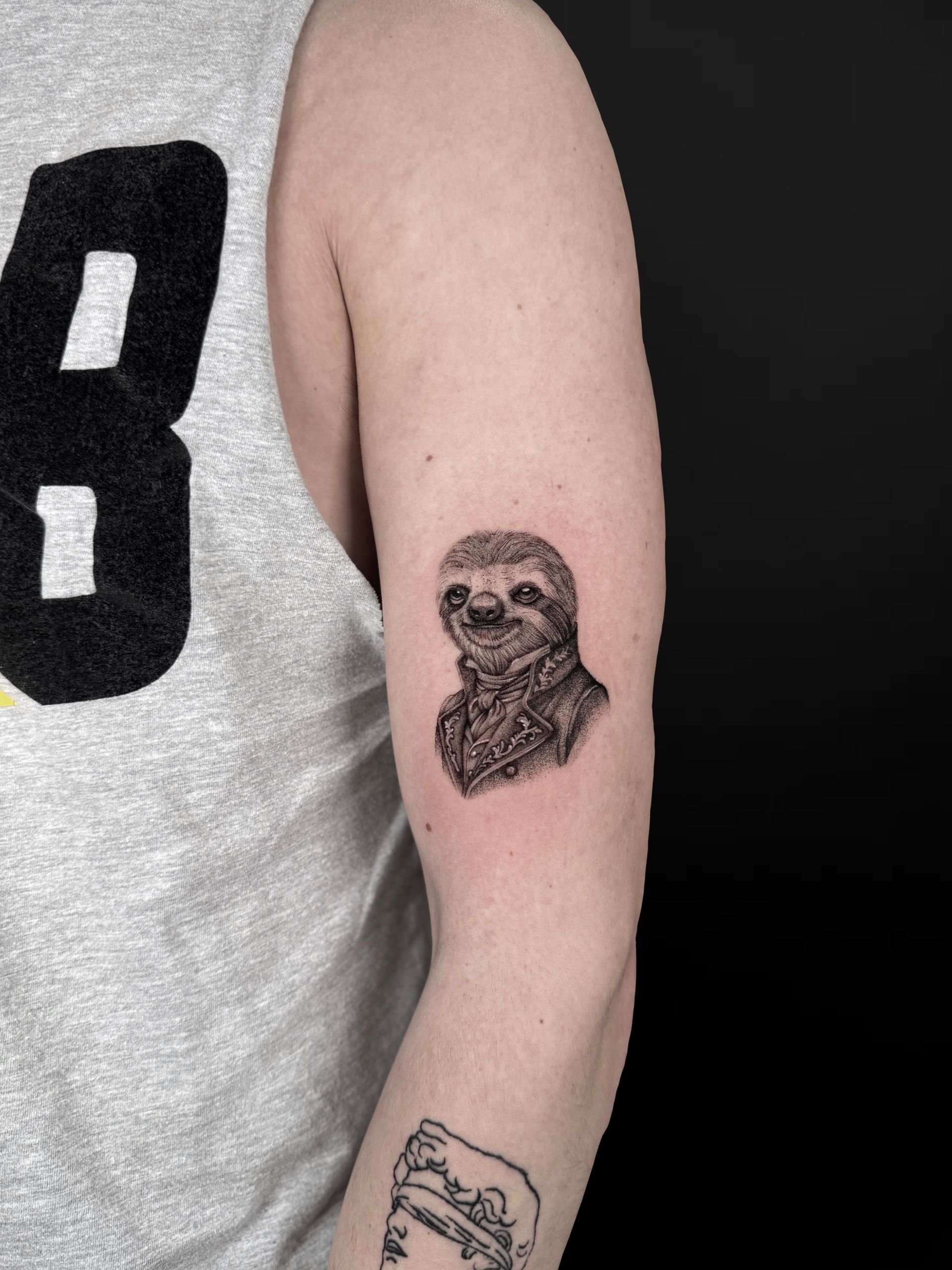A detailed black-ink tattoo of a sloth dressed in a formal Victorian-style coat on a person's upper arm.