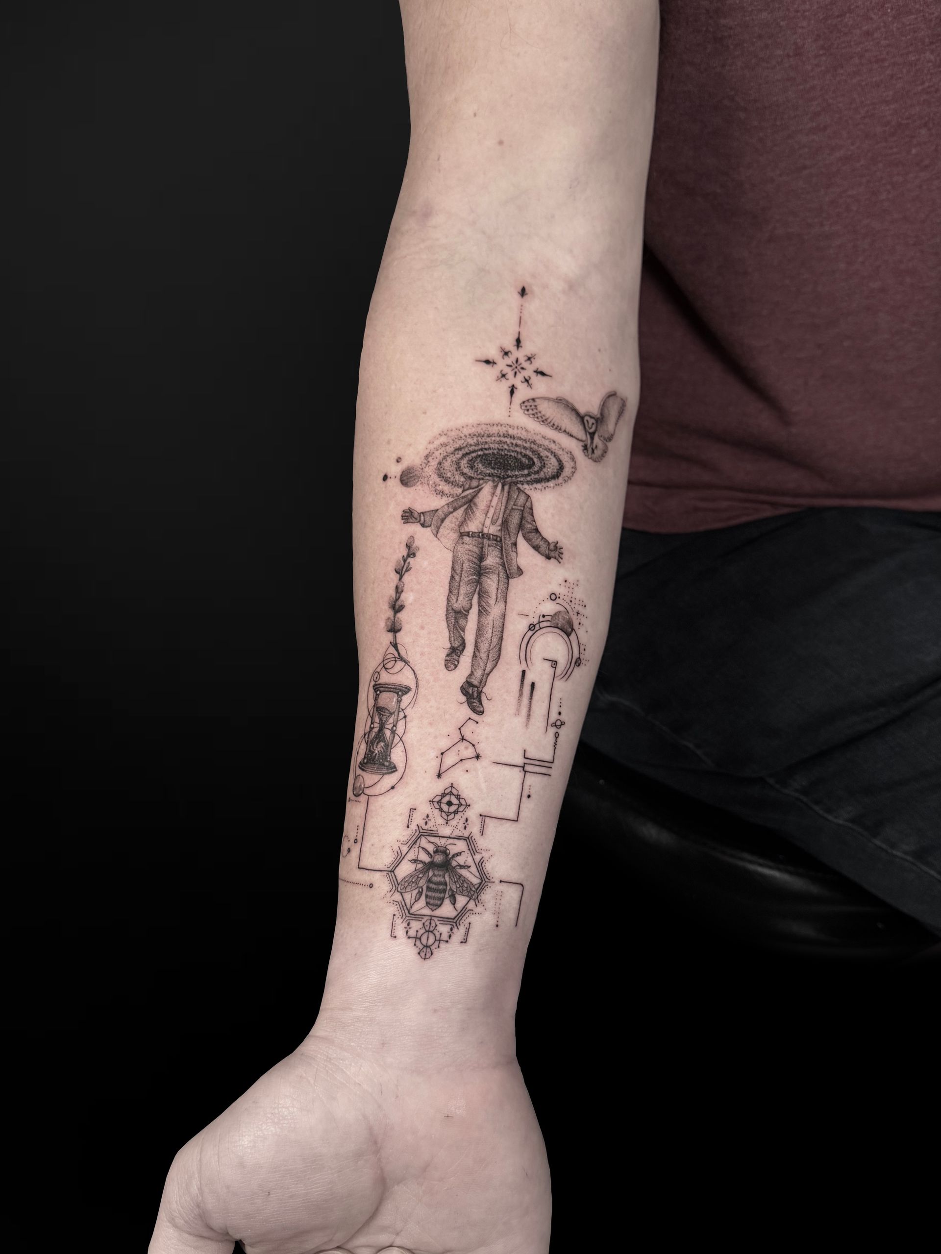 A black-ink forearm tattoo featuring a floating figure with a galaxy head, a compass, an hourglass, and an owl.