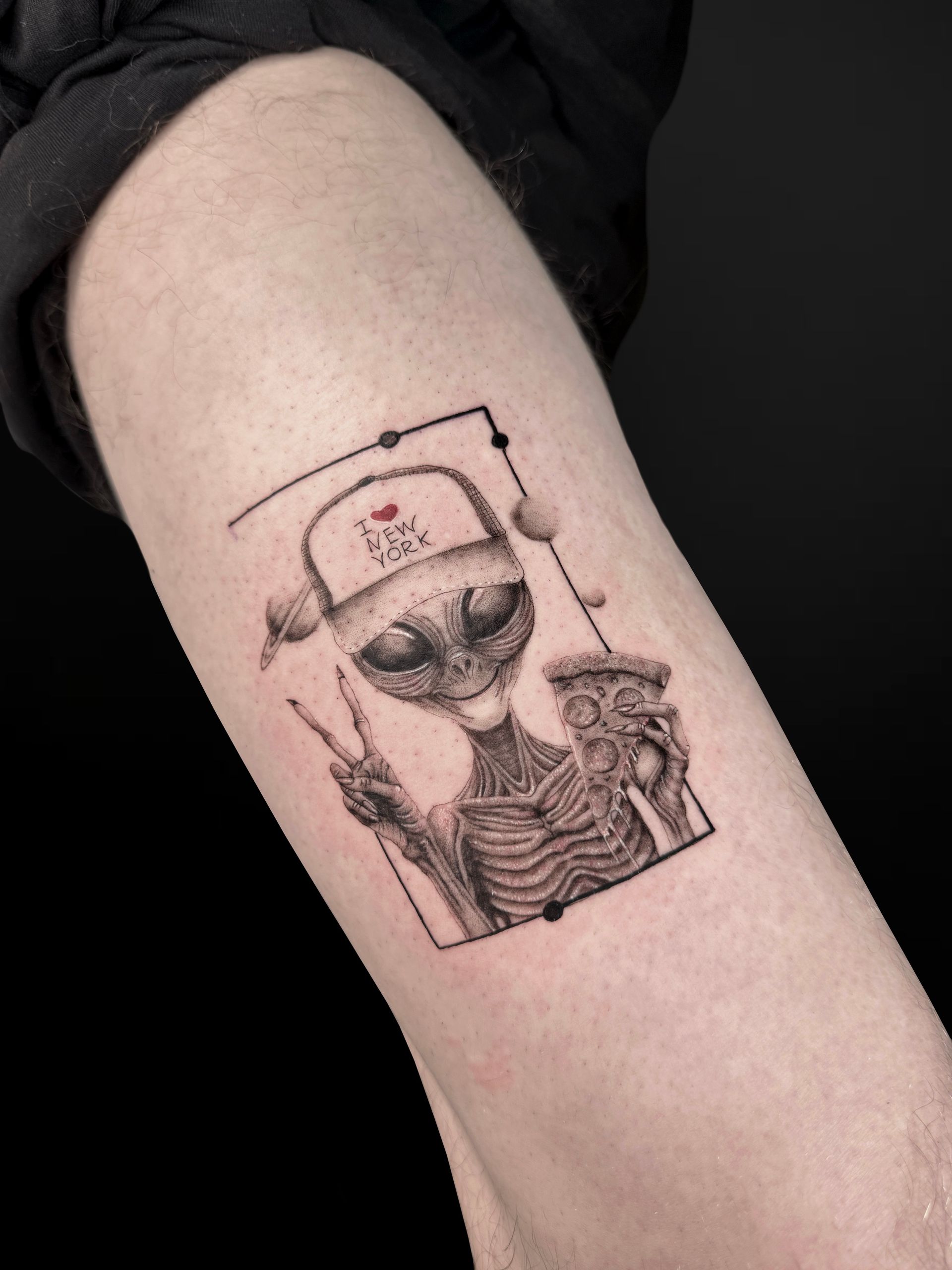 A fine-line tattoo on skin of a skeleton alien wearing a baseball cap, making a peace sign and holding a slice of pizza.