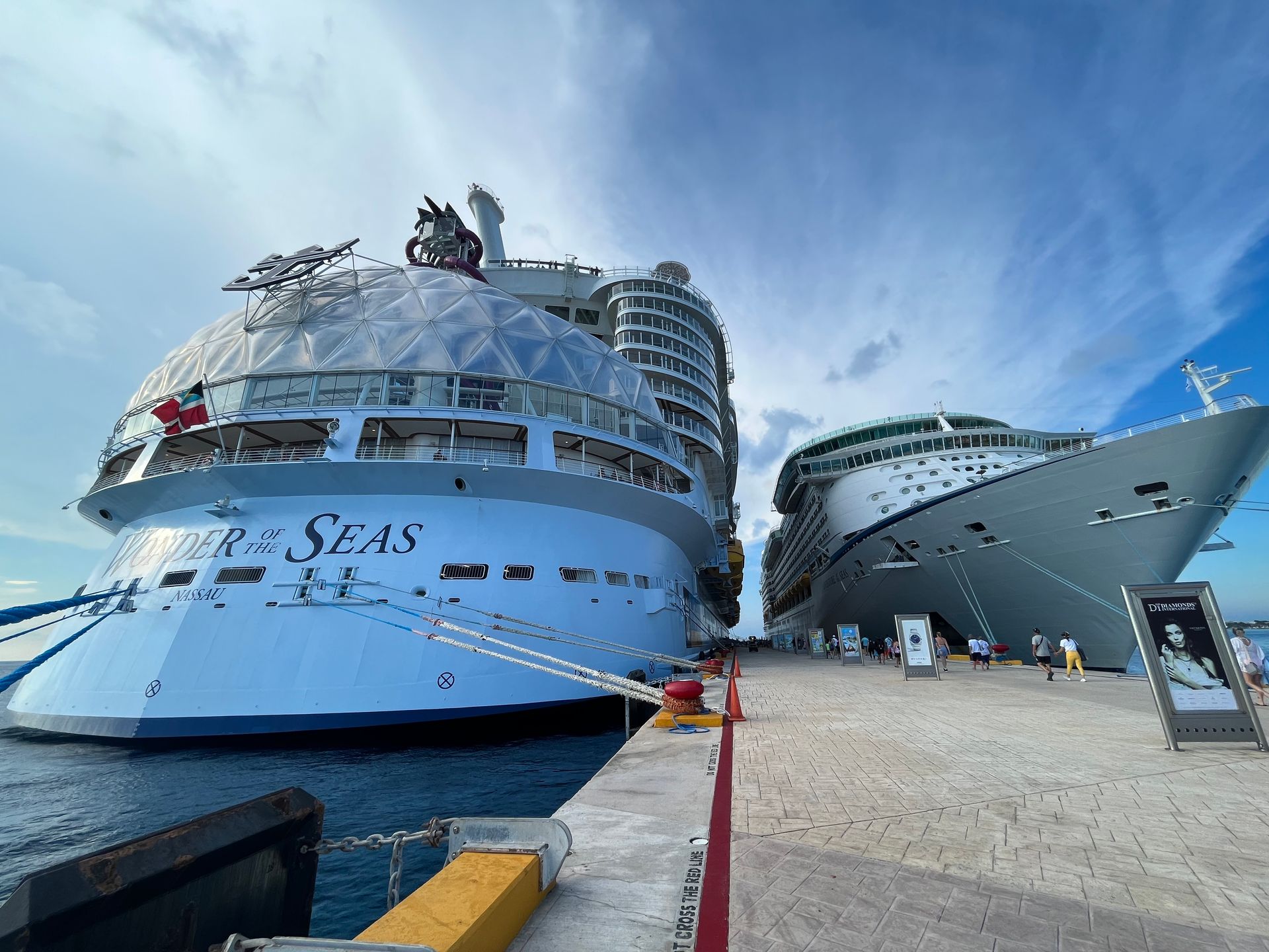 Two cruise ships docked side-by-side at a pier, under a partly cloudy sky. The larger ship is blue and white.