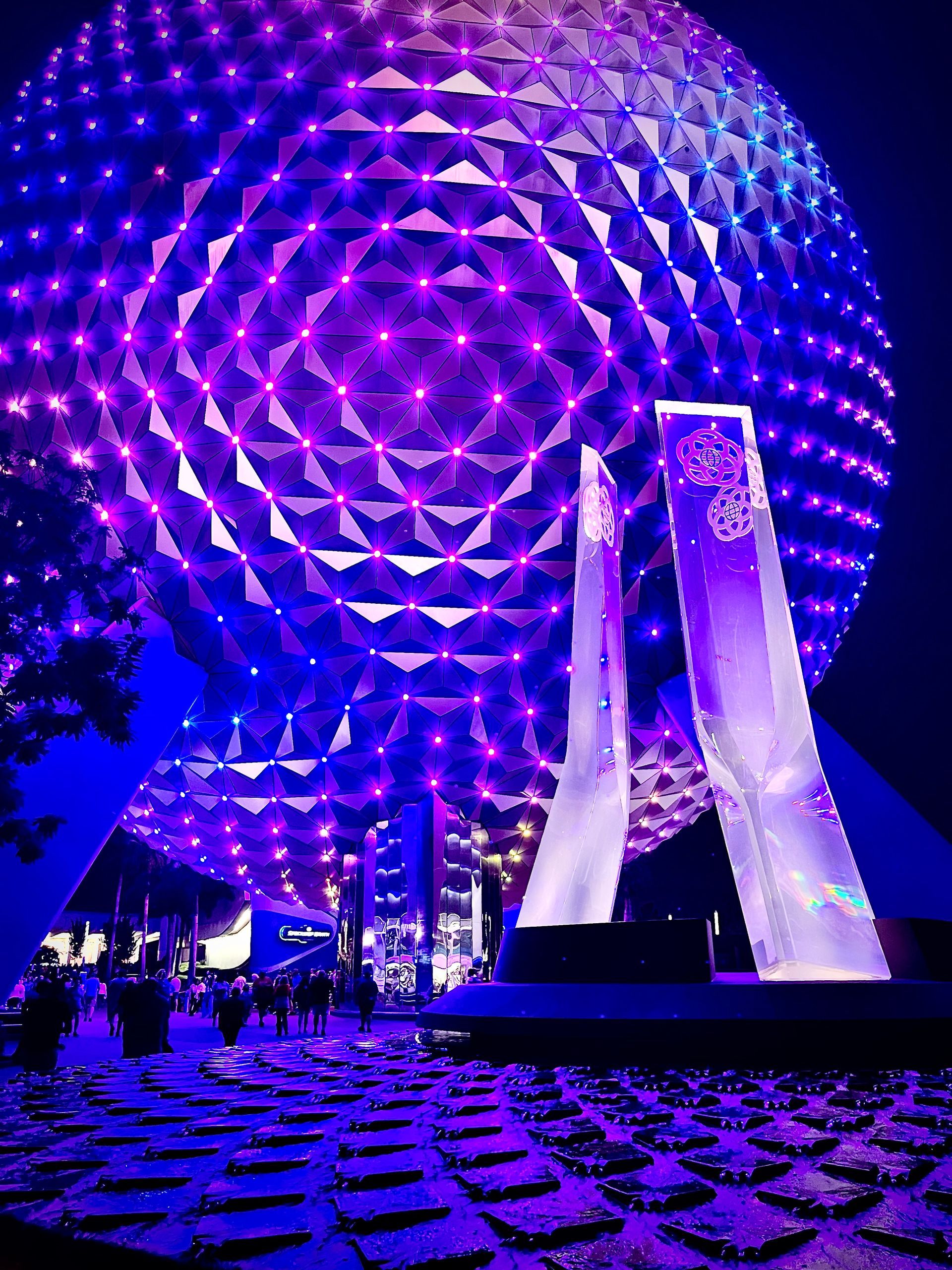 Illuminated geodesic dome at night with purple lights and a modern sculpture in front
