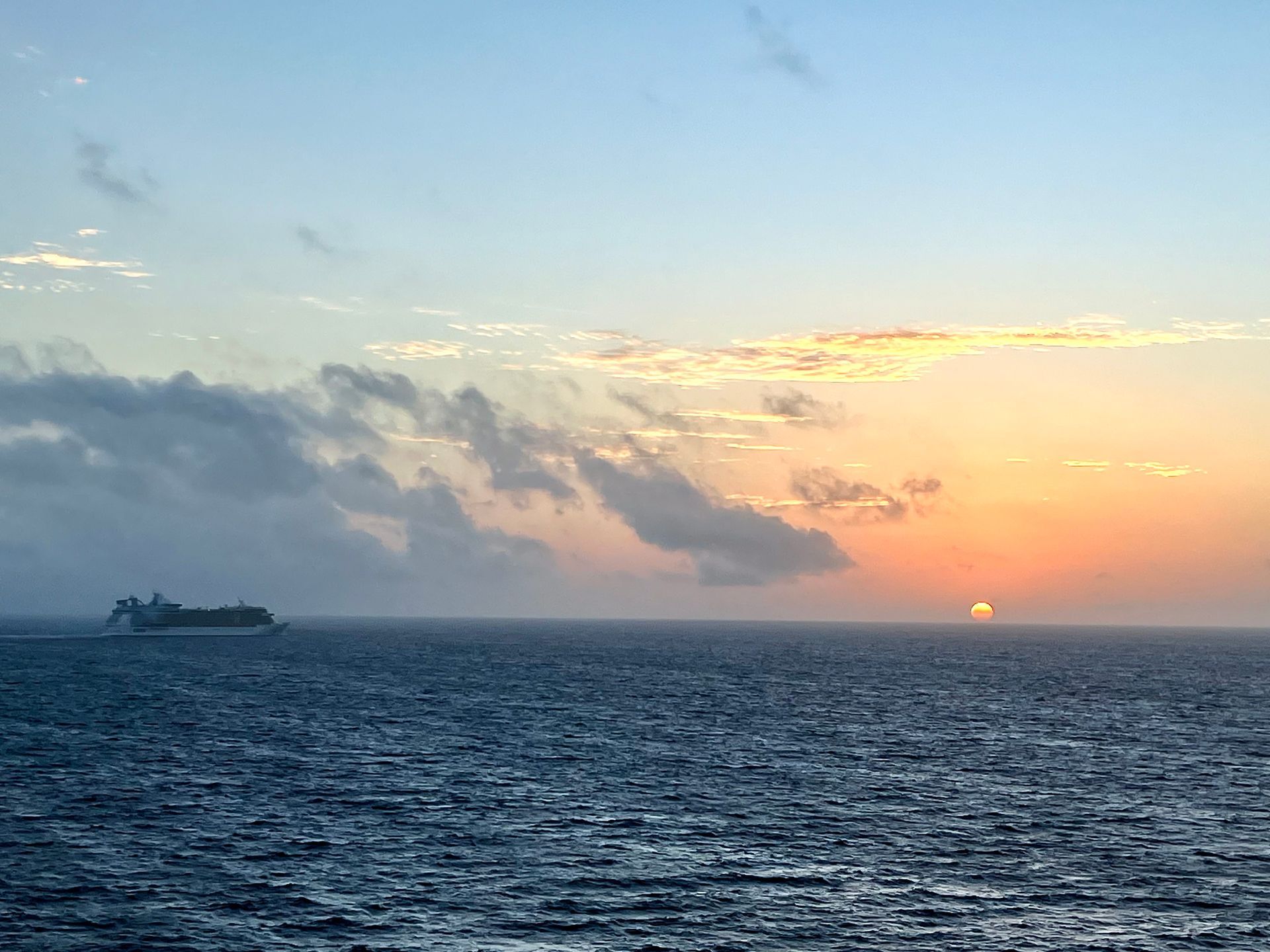 Sunset over ocean, cruise ship on the left, orange and yellow sky, clouds.