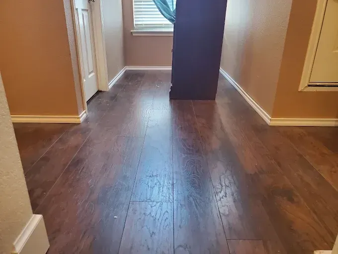 A hallway with dark wood-look laminate flooring leading toward a window with blue curtains and a black object.
