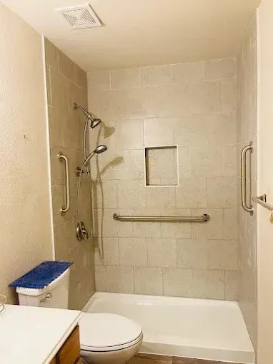 A white shower base and tiled walls with three metal grab bars, a handheld showerhead, and a small recessed shelf.