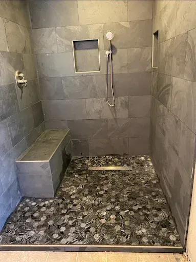A walk-in shower with gray tile walls, a built-in bench, a pebble floor, a handheld showerhead, and two recessed niches.