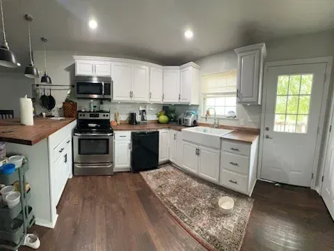 A kitchen with white cabinets, dark wood countertops, stainless steel appliances, and a patterned rug on a wood floor.