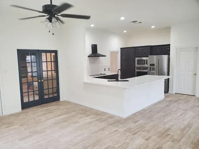 A modern kitchen with white counters, dark cabinetry, and a ceiling fan, opening into a room with French doors.