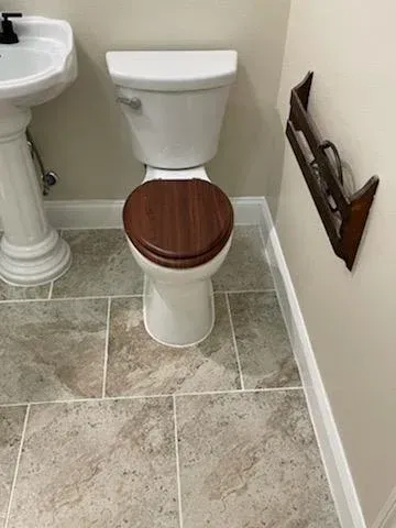 A bathroom with a white toilet featuring a dark wood-toned seat, adjacent to a pedestal sink and a wall-mounted wood rack.