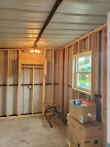 Interior view of a shed under construction, showing wooden wall framing, an installed window, and a ceiling light fixture.