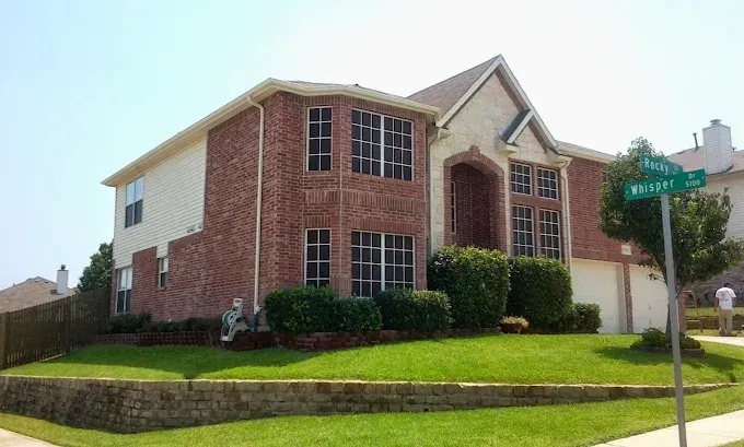 A two-story brick and siding house with a green lawn, a stone retaining wall, and a street sign on a sunny day.
