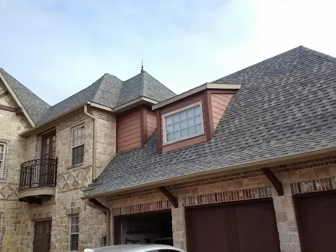 A stone house exterior with a multi-gabled shingled roof, a dormer window, and an attached garage under a blue sky.