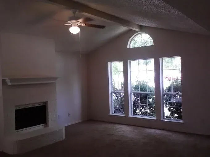 A living room with a white fireplace, vaulted ceiling, ceiling fan, and three tall windows topped by a semi-circle window.