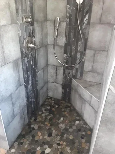 A walk-in shower with grey tiled walls, dark slate accents, a built-in corner seat, and a river rock pebble floor.