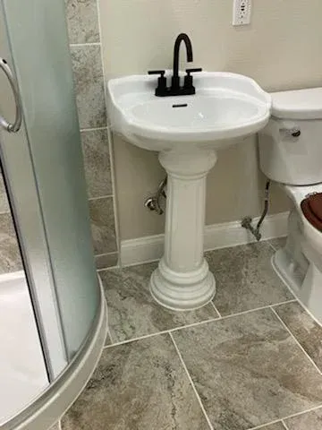 White pedestal sink with a matte black faucet in a bathroom with beige tile floors and a glass-enclosed shower.