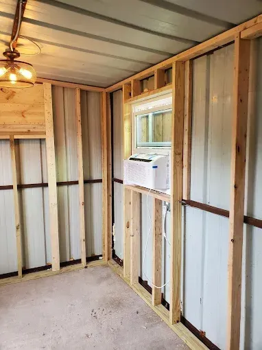 Interior view of a storage shed under construction with wooden wall studs, a window AC unit, and a hanging light fixture.