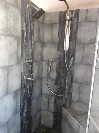 A modern walk-in shower with charcoal-colored tile walls, a rain shower head, handheld sprayer, and a corner seat.