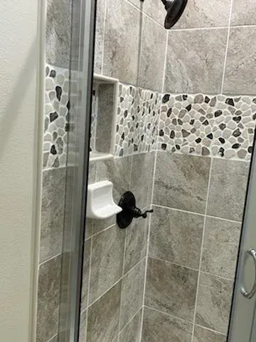 A shower interior with neutral stone-look square tiles, a pebble-style accent border, a recessed niche, and black fixtures.