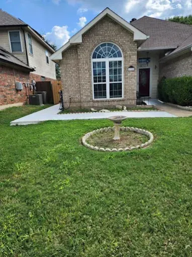 A brick home with a front lawn, featuring a circular birdbath in the center and a new concrete walkway near the entrance.
