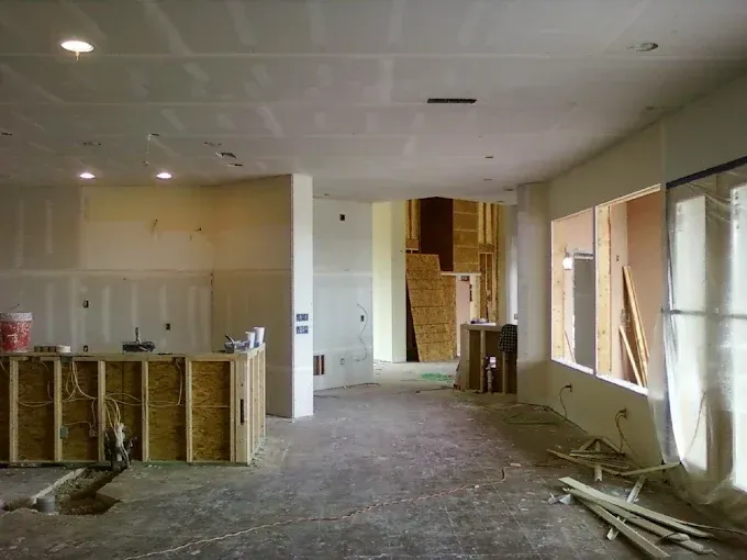 An interior room under construction with drywall, a wooden framed island, exposed wooden structure, and large windows.