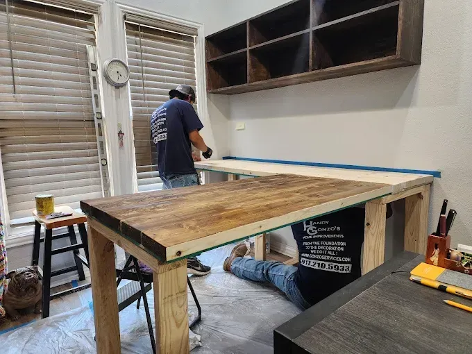 Two workers assemble a wooden desk in a room with a wall-mounted shelf, window blinds, and painting supplies.