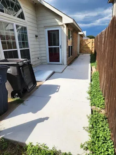 A concrete path leads along the side of a house toward a wooden gate, flanked by a trash bin and a wooden fence.