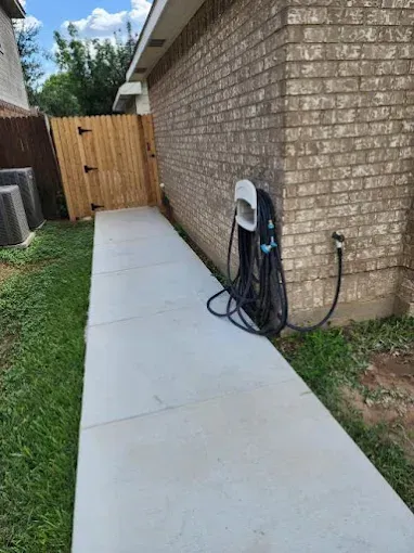 A straight concrete sidewalk leads to a wooden fence next to a brick house exterior with a hose on a reel.