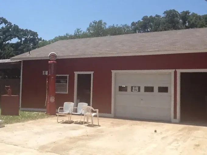 A red detached garage with white trim, a vintage gas pump, and three white patio chairs on a concrete driveway.