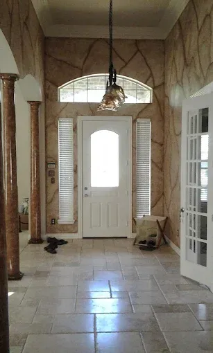 A home entryway with stone-patterned walls, two columns, a white front door with sidelights, and light-colored tile floors.