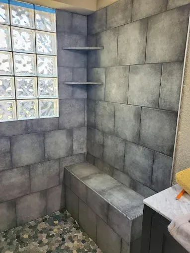 A shower stall featuring grey, square-tiled walls, a built-in bench, two corner shelves, and a glass block window.