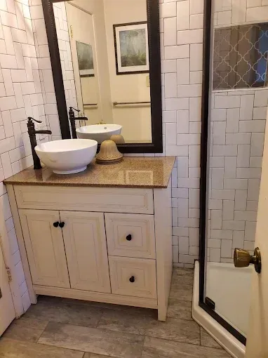 A bathroom vanity with a white vessel sink, dark faucet, beige countertop, and white cabinetry, next to a glass shower.