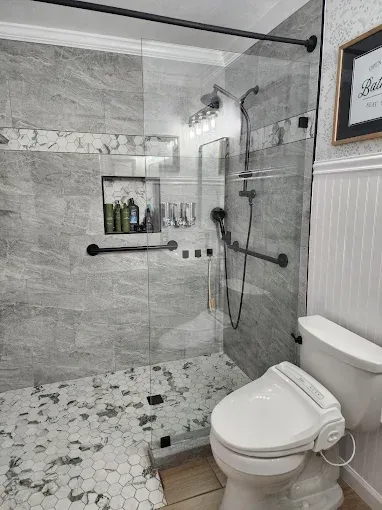 Modern bathroom with a glass-enclosed walk-in shower, grey stone tile, pebble floor, grab bars, and a white toilet.