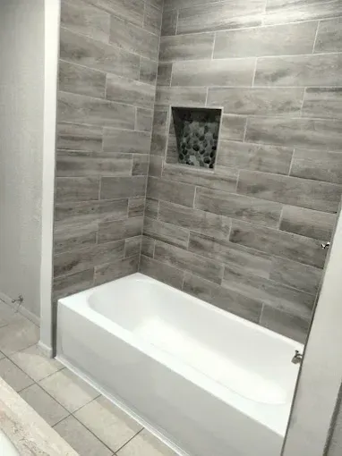 A bathroom shower featuring a white bathtub and grey, wood-look tiled walls with a small recessed shelf.