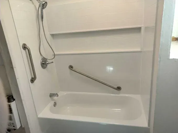A white bathtub shower enclosure featuring a handheld showerhead, two safety grab bars, and integrated storage shelves.