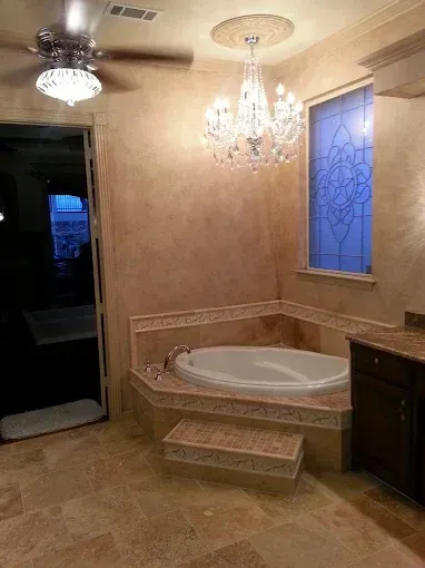 A corner bathtub with beige stone tile surround and steps, under a crystal chandelier, next to a blue stained glass window.