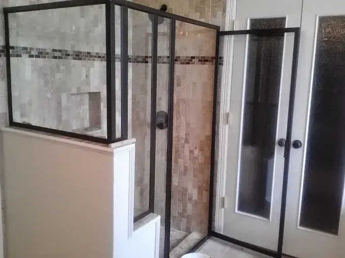 A modern shower enclosure with black metal frames set against tan stone tiled walls, next to a door with glass panels.