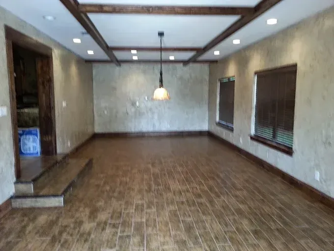 A spacious room with wood-look tile floors, textured light walls, and a wood-beamed ceiling with a hanging pendant light.