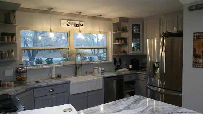 Modern kitchen with gray cabinets, a white farmhouse sink, stainless steel appliances, and a marble-patterned island.