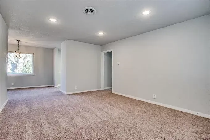 A spacious room with beige carpet, light gray walls, recessed lighting, and a view into a secondary room with a window.